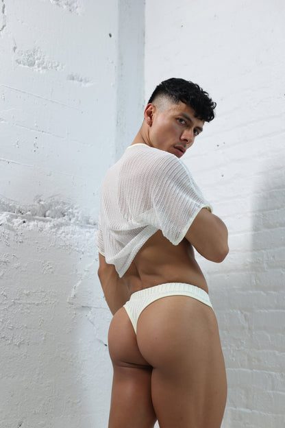 Fabian Divani modeling a sheer white mesh crop top and swim thong set—handmade men's swimwear—posed in front of a white brick wall