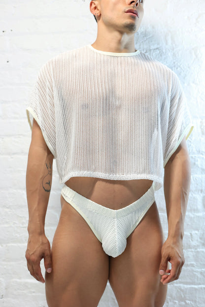 Fabian Divani modeling a sheer white mesh crop top and swim thong set—handmade men's swimwear—posed in front of a white brick wall